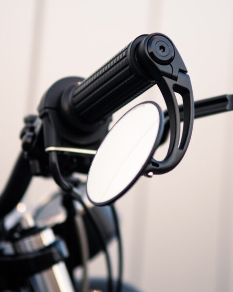 Close-up of a CNC-engineered motorcycle handlebar end mirror mounted on black handlebars, with a clear reflective surface, showing part of the motorcycle's grip and control area.